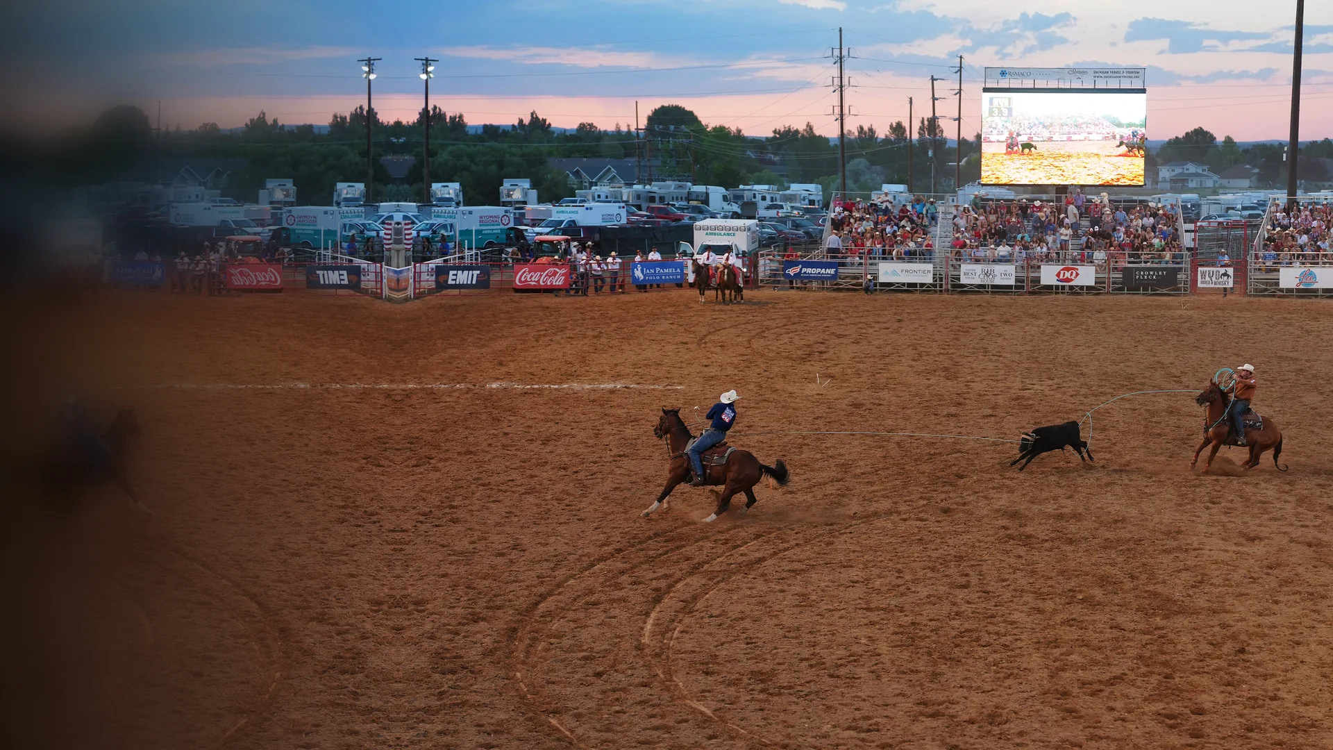 Two riders on horseback roping a running steer across a dirt rodeo arena at dusk, with a crowd in the stands and a big screen above the fence.