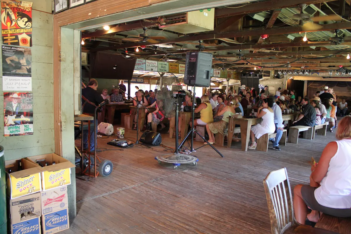 People seated at wooden tables in a large open-sided wooden dance hall while a band plays on a small stage